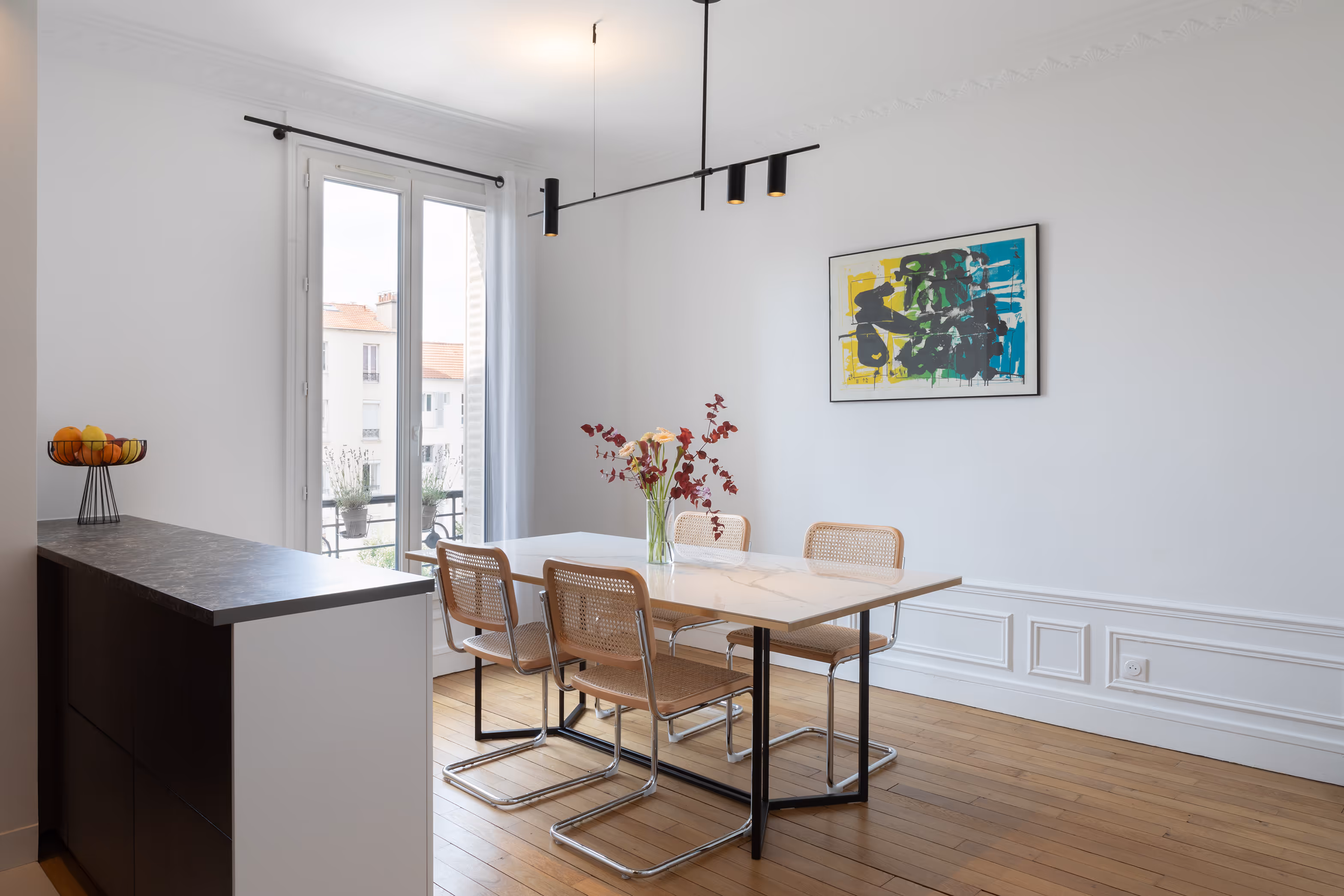 Salle à manger minimaliste avec table rectangulaire blanche, quatre chaises en cannage, vase avec fleurs, suspension noire, et grande fenêtre donnant sur des bâtiments. Projet de rénovation Daumesnil à Paris 12.