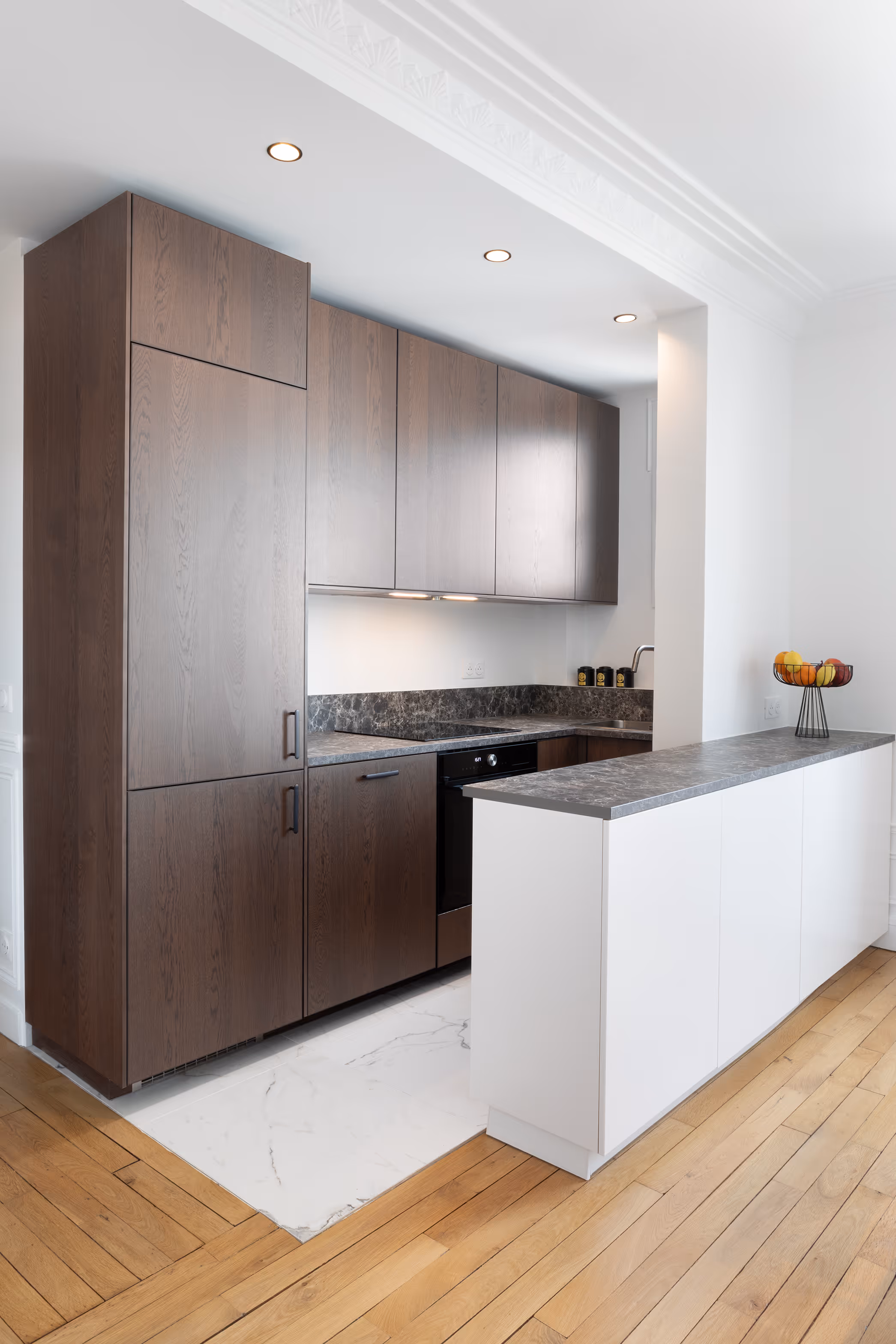 Cuisine contemporaine sur mesure en chêne foncé avec plan de travail en pierre grise et îlot blanc avec corbeille de fruits. Projet de rénovation Daumesnil à Paris 12.
