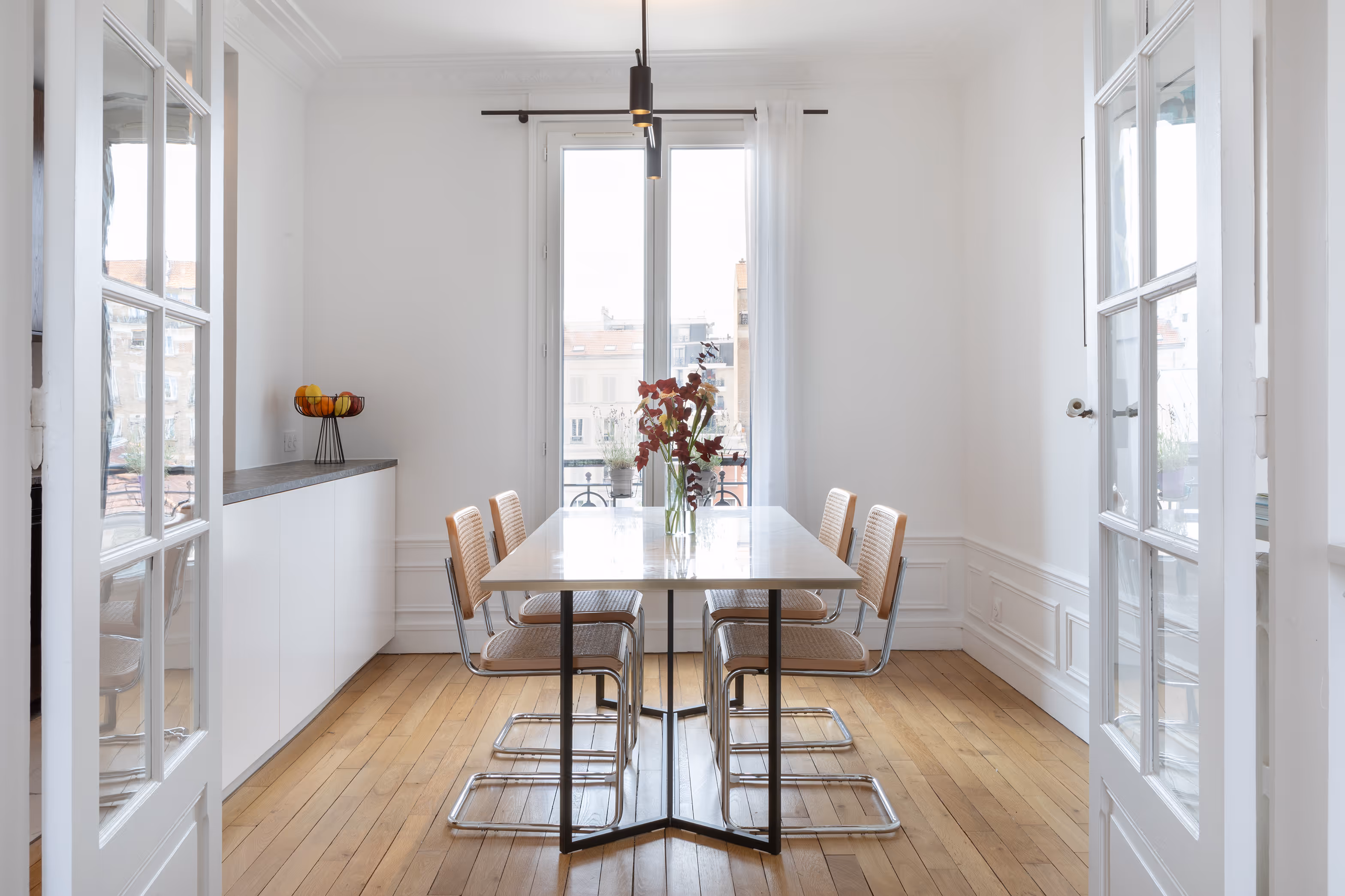Salle à manger moderne et épurée avec table blanche, quatre chaises en rotin et plateau en verre, parquet en bois clair, et baie vitrée donnant sur un balcon urbain. Projet de rénovation Daumesnil à Paris 12.