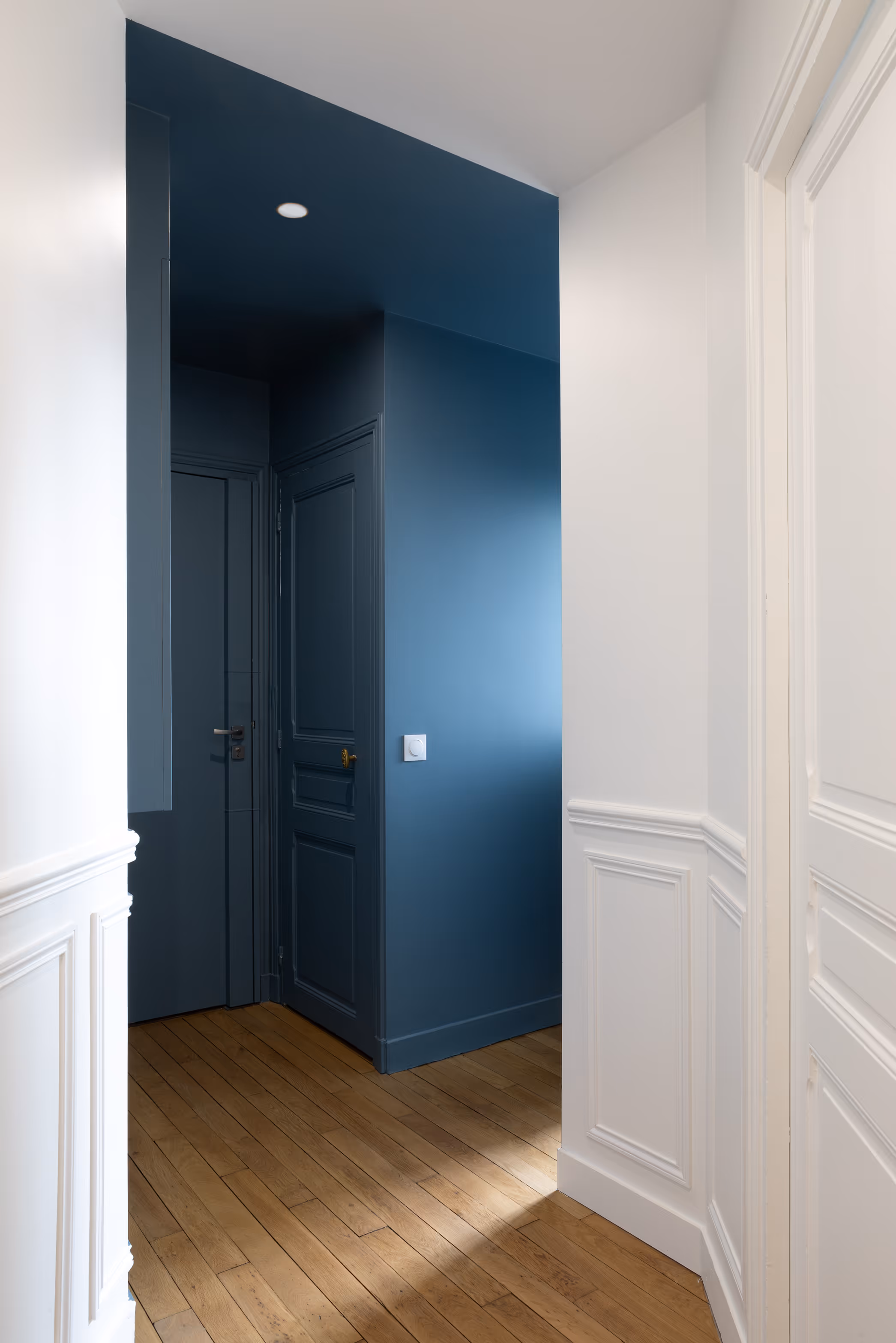 Aménagement de couloir avec murs blancs et sol en parquet clair, structuré par un mur bleu pétrole et des portes intégrées assorties. Rénovation d'un appartement haussmannien à Paris.